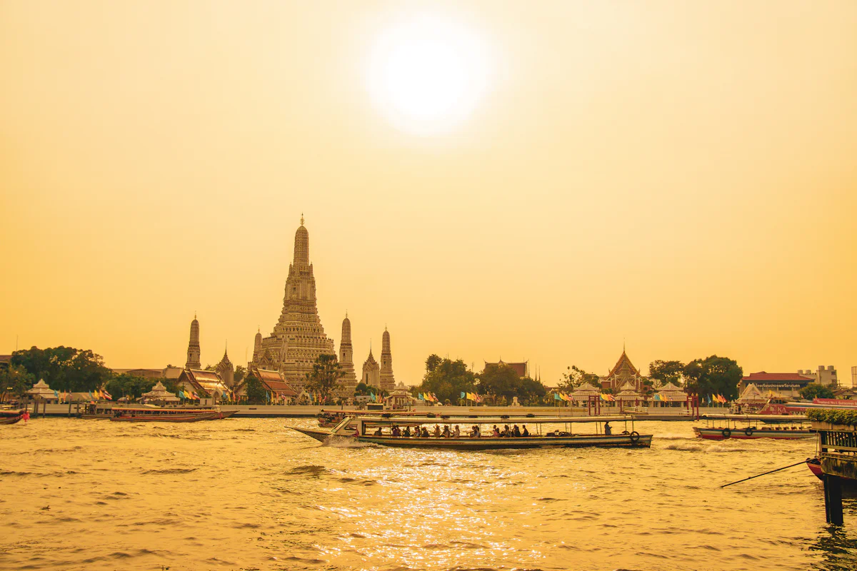 Wat Arun on the Chao Phraya river in Bangkok at golden hour, with passenger boats on the water