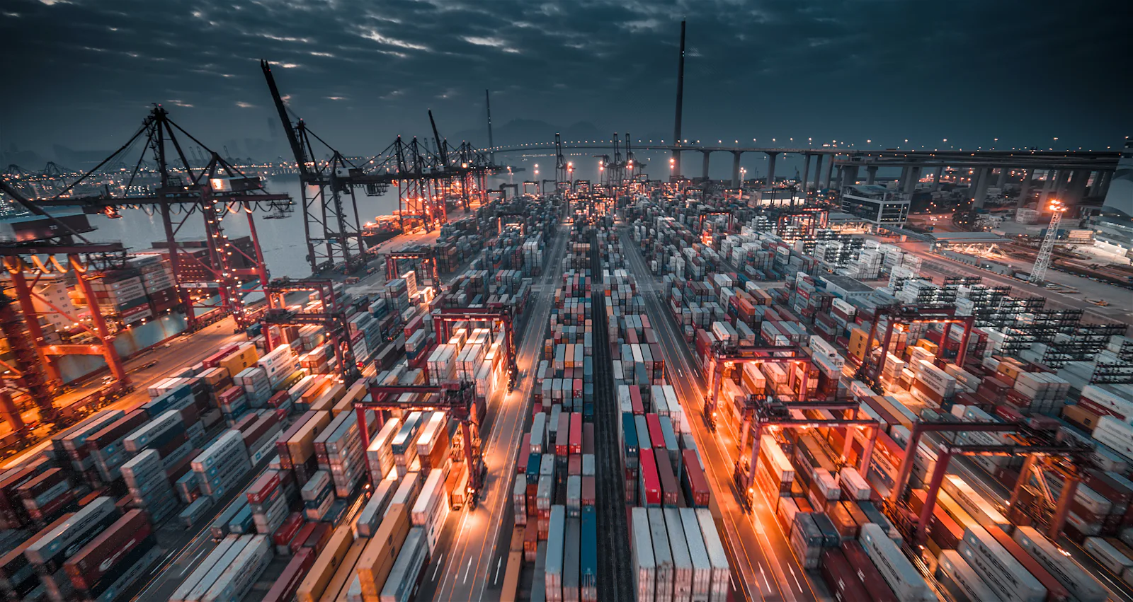 A container terminal at night, rows of stacked containers lit from above with gantry cranes along the quay